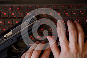 A gun lies on a black laptop keyboard with red backlight and a woman& x27;s hand on the keyboard, security threat, cyber