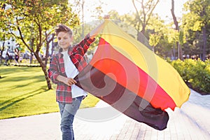Guy of 9 years old riding skateboard on sunny road in the evening in the city park. Holding fluttering flag of Germany. Happy