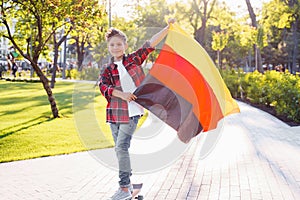 Guy of 9 years old riding skateboard on sunny road in the evening in the city park. Holding fluttering flag of Germany. Happy