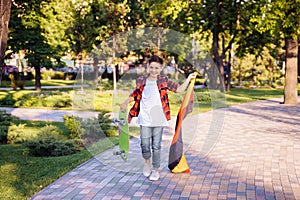 Guy of 9 years old riding skateboard on sunny road in the evening in the city park. Holding fluttering flag of Germany. Happy