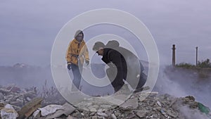 Guy assisting to Girl to jump between piles of Garbage. Couple of survivors with weapons in the smoky and unkind