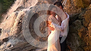 A guy attracts a girl standing among the rocks under the waterfall during the sunset, slow motion