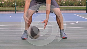 A guy basketball player dribbling ball between his legs at outdoor court. An athlete in a gray sports uniform hits the