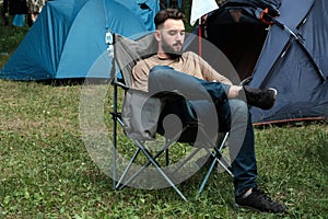 A guy with a beard in jeans and a t-shirt is sitting on a folding chair near the tents. The concept of man`s leisure in