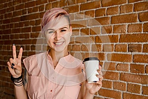 Young beautiful smiling girl holds paper cup of coffee on a brick wall background