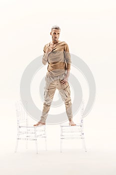 A guy in a beige jumper and cargo pants stands on transparent chairs on a white background