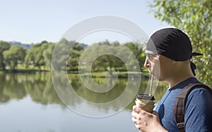 A guy in a black bandana and sunglasses drinks coffee from a paper cup through a straw while walking along the pond