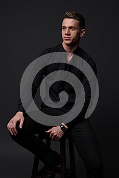 a guy in a black shirt and black pants sits on a dark chair, on a gray background