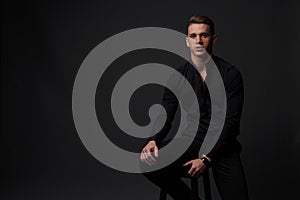 a guy in a black shirt and black pants sits on a dark chair, on a gray background