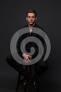 a guy in a black shirt and black pants sits on a dark chair, on a gray background