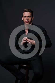 a guy in a black shirt and black pants sits on a dark chair, on a gray background