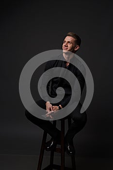 a guy in a black shirt and black pants sits on a dark chair, on a gray background