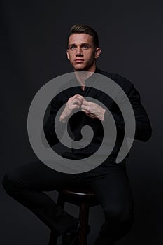 a guy in a black shirt and black pants sits on a dark chair, on a gray background