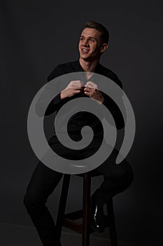 a guy in a black shirt and black pants sits on a dark chair, on a gray background