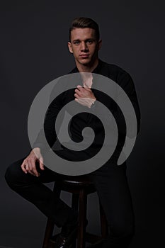 a guy in a black shirt and black pants sits on a dark chair, on a gray background