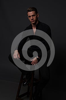 a guy in a black shirt and black pants sits on a dark chair, on a gray background