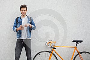 The guy in a blue denim jacket standing on wall background. young man near orange bicycle. Smiling student with bag