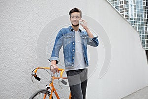 The guy in a blue denim jacket standing on wall background. young man near orange bicycle. Smiling student with bag