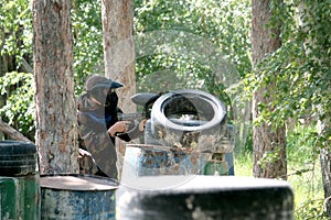 A guy in camouflage clothes from a blue team with weapons in his hands peeks out from behind an iron barrel and tires. Sport game