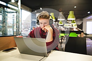 Guy in casual clothing and headphones, sitting at the table in a cafe, working on a laptop, focused on the screen