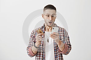 Guy checks his bank account via smartphone. Portrait of shocked good-looking man in glasses holding credit card and