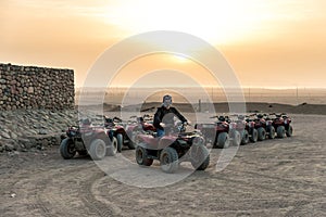 A guy with a closed handkerchief on his face sits on a quad bike against the backdrop of the sun rising over the desert