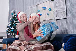 The guy closes the girl`s eyes, while the girl is holding a Christmas gift box. . Indoors.