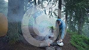 A guy cooks food in a cauldron on a fire on a camping trip with a tent.