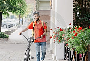 Guy curier with beard with backpack in protective helmet, holds bicycle and walks down the street