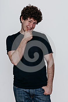 a guy with curly hair in a black T-shirt and jeans smiles at the camera. Hand near the chin.