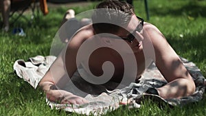 Guy in dark glasses lies on lawn, on mat, looks camera, smiles, jokes, laughs