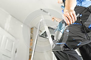 Guy in work clothes and with an electric screwdriver in his hand is standing on the stairs. Against the background of renovation