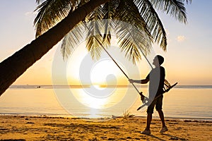 The guy enjoys the sunset riding on a swing on the ptropical beach. Silhouettes of a guy on a swing hanging on a palm tree,