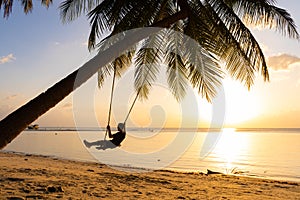 The guy enjoys the sunset riding on a swing on the ptropical beach. Silhouettes of a guy on a swing hanging on a palm tree,