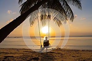 The guy enjoys the sunset riding on a swing on the ptropical beach. Silhouettes of a guy on a swing hanging on a palm tree,