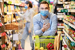 Guy in face mask with smartphone shopping in hypermarket