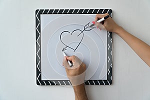 A guy and a girl draw a heart with markers on a white board. A mask and a female hand against a white board with a symbol of love