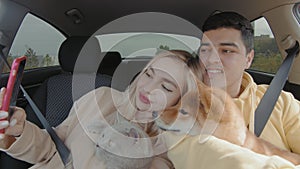 A guy and a girl are driving in a car with a cat and a dog. A guy and a girl take a selfie on their phone in the car