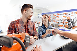 The guy and the girl pay for the purchase of a chainsaw.