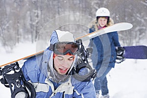 A guy and a girl in snowboarding clothes are walking along the ski slope. They are holding a snowboard.