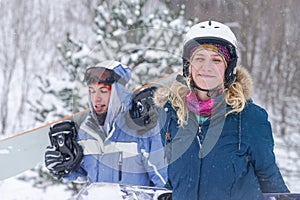 A guy and a girl in snowboarding clothes are walking along the ski slope. They are holding a snowboard.