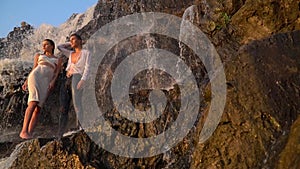 The guy and the girl are standing by the waterfall and watching the sunset