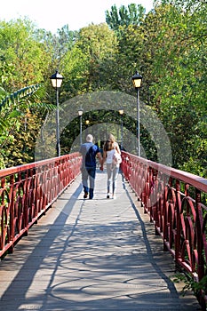 A guy and a girl walk holding hands with their backs turned on a pedestrian bridge with red railings
