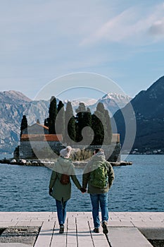 Guy and girl are walking along the pier, looking at the mountains behind the island of St. George. Montenegro. Back view