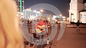 A guy with a guitar playing in the town square at night Visible city lights.