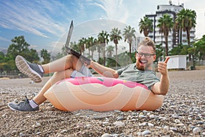 Guy happy smile in glasses works at a laptop while sitting in an inflatable circle in the form of donut on beach with palm trees