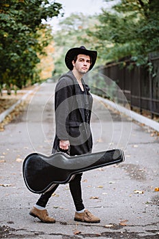 A guy in a hat and a guitar walking down the street.
