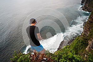 Guy in headphones stands on a cliff by the ocean at sunset in bali