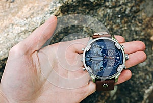 Guy is holding fashionable elegant men`s watch in hands. Stylish accessories in the forest on stone. Copy space place