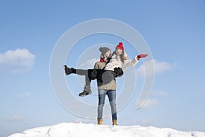 Guy holds girl in his arms on snow-covered hill against blue sky. Winter sunny day in mountance